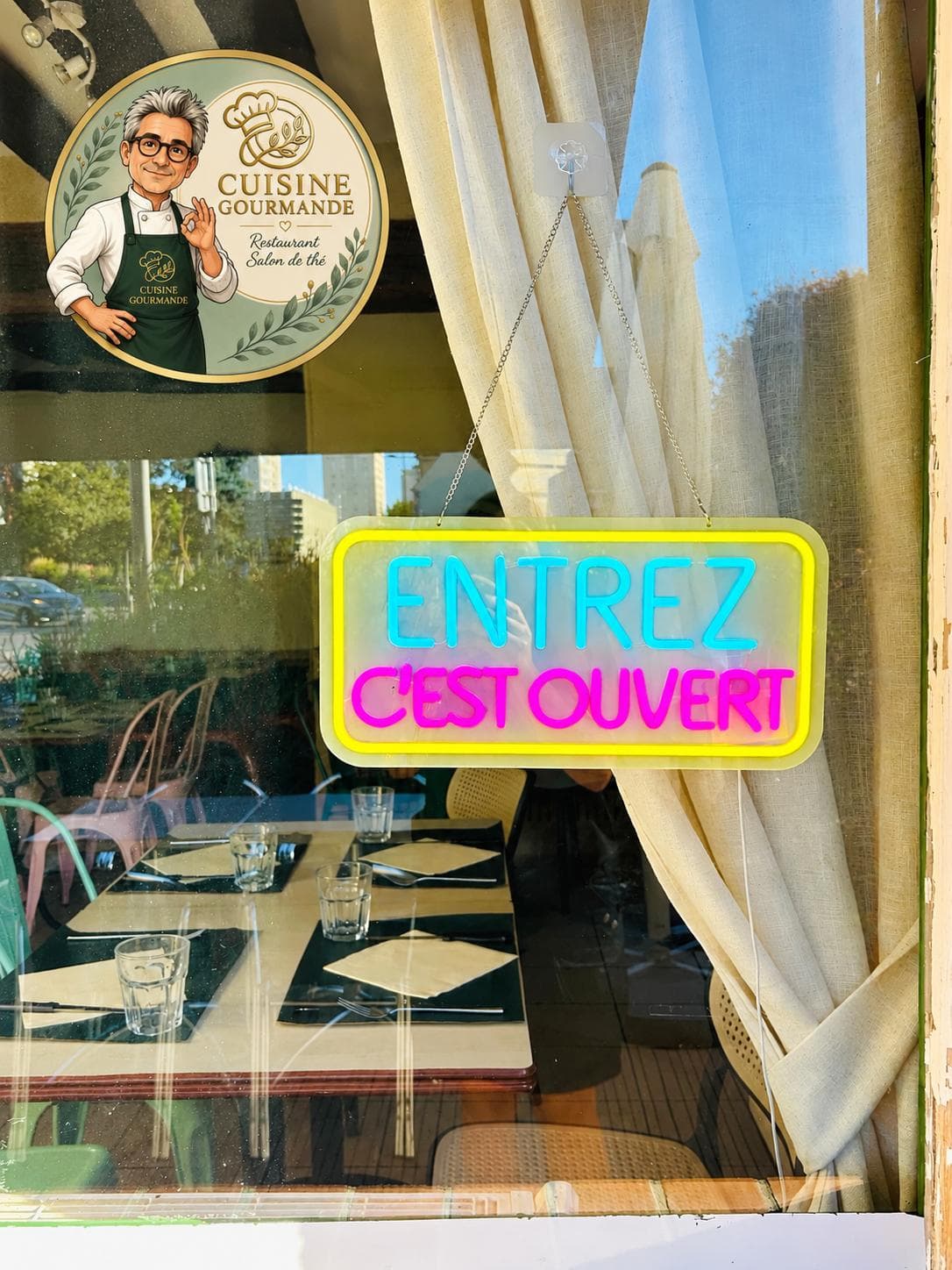 Vitrine du restaurant avec le logo et l'enseigne « Entrez, c'est ouvert », tables dressées en salle